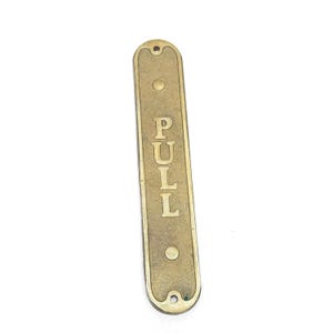 May include: A brass door pull sign with the word "PULL" engraved in a vertical orientation.