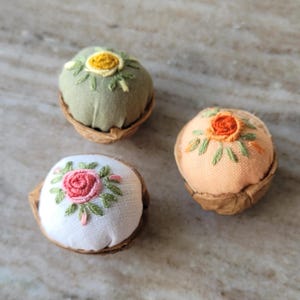 以下が含まれることがあります： クルミの殻で作られた3つの小さなピンクッション。それぞれに花柄が刺繍された布製のドームが入っています。1つはセージグリーンの布地に黄色い花、もう1つは白地にピンクのバラ、3つ目はピーチ色の布地にオレンジ色の花が刺繍されています。