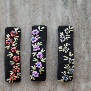 以下が含まれることがあります： 黒い長方形のヘアバレッタ3つ。それぞれに花の刺繍が施されています。各バレッタは異なる配色で、赤とオレンジ、紫と青、銀と緑です。花は小さなビーズでアクセントがつけられています。