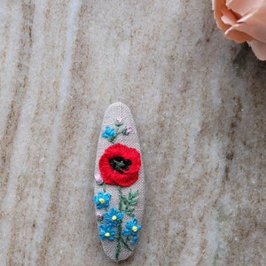 May include: A gray linen hair clip with a red poppy and blue forget-me-nots embroidered on it.
