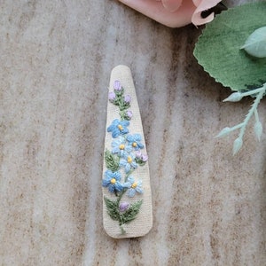 以下が含まれることがあります： ベージュ色の生地に、青と紫の花と緑の葉を刺繍したヘアクリップ。 花柄のデザインはクリップの中央に配置され、上部が丸く、わずかに先細りになっています。 手作りのアクセサリーです。