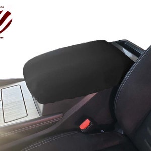 May include: Black center console cover in a car interior. The cover is made of a soft material and fits over the armrest. The image includes the text "Made in USA" and "With Pride" with an American flag heart.
