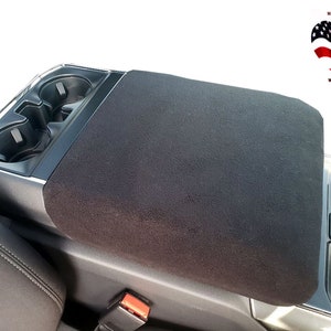 May include: Black centre console armrest cover for a car. The cover is made of a soft, plush fabric and has two cup holders.