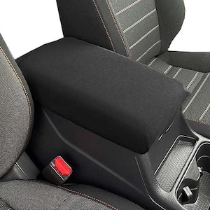 May include: A black fabric center console armrest cover installed in a vehicle. The cover features a smooth, solid black surface that fits over the center console lid, positioned between two car seats with grey and black textured upholstery.