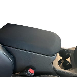 May include: A black fabric car center console armrest cover featuring a smooth, matte texture that fits over the top of the console lid, positioned between the front vehicle seats near the cup holders and a red seatbelt release button.