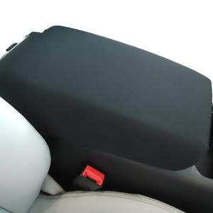 May include: Black car seat armrest cover with a red pull tab. The cover is made of a soft, black fabric.