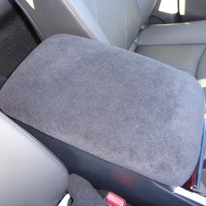 May include: A gray car armrest with a soft, plush fabric covering. The armrest has a built-in cup holder.