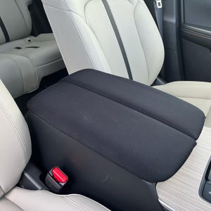 May include: A black padded car armrest is positioned between two cream-colored car seats. The armrest is rectangular with a textured surface. A red seatbelt buckle is visible. The car interior features wood grain and black accents.