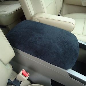 May include: A navy blue, plush center console cover in a car interior. The cover is rectangular with rounded edges, designed to fit over the armrest. The surrounding interior features beige leather seats and trim.