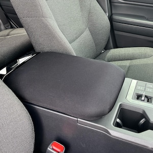 May include: Black center console cover in a car interior. The cover is made of a soft, dark fabric and fits over the armrest. The car seats are gray, and the console has various buttons and cup holders.