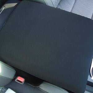 May include: Black car seat cover with a smooth, textured surface. The cover is designed to fit over the center console armrest.