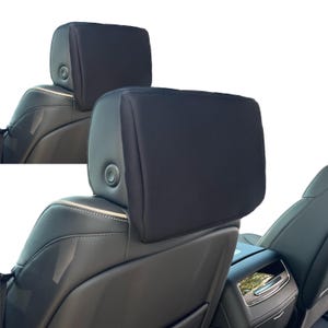 May include: Two black car headrests are shown, attached to the seats. The headrests are rectangular and appear to be made of a soft, dark fabric. The seats are black leather with visible stitching and a silver accent.