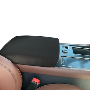 May include: Black padded center console cover in a car interior. The cover is rectangular and fits over the armrest. The car interior features brown leather seats and a silver console with various controls and storage compartments.