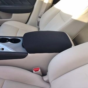 May include: Black car armrest cover with a black fabric material. The cover is installed on the center console of a car.