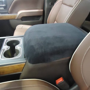 May include: Black suede center console armrest cover for a car. The cover is installed over the existing armrest and has a smooth, soft surface.
