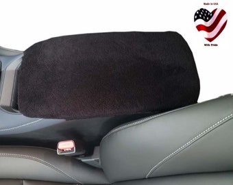 May include: A black, plush center console cover is shown, designed to fit over a car's armrest. The cover is rectangular and appears to be made of a soft, textured material. The image includes a car seat and the text "Made in USA" with a heart-shaped American flag.