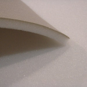 May include: Close-up of a beige fabric folded over a white foam material. The fabric has a smooth texture, while the foam has a slightly textured surface.