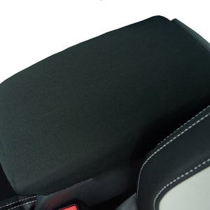 May include: Black armrest cover with a smooth, textured surface. The cover is designed to fit over a car's center console armrest.