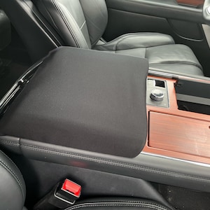 May include: Black padded center console armrest cover in a car interior. The cover is rectangular and fits over the console. The interior features black leather seats, wood trim, and chrome accents.