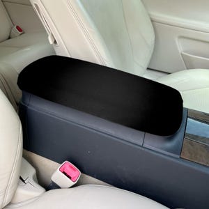 May include: A black padded console cover is placed on the center console of a car. The cover is rectangular and fits over the armrest. The car's interior features light-colored leather seats and a dark gray console.