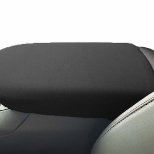 May include: A black car headrest, with a rounded rectangular shape, is positioned in front of a car dashboard and a gray leather seat. The headrest is made of a soft, textured material.