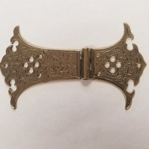 May include: A gold-toned metal hinge with intricate floral designs and a decorative, ornate finish. The hinge is closed and has a detailed, intricate design.