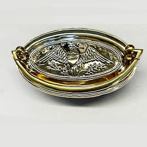 May include: A gold-toned oval cabinet pull with a raised, silver-toned eagle design. The eagle has its wings spread and is holding a branch in its talons.
