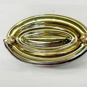 May include: A gold oval-shaped cabinet pull with a raised, concentric ring design. The pull has a smooth, polished finish and two small, round knobs on either side.