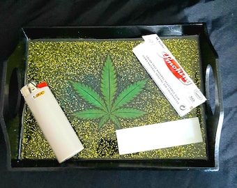 May include: A black tray with a green marijuana leaf design, a white lighter, a pack of Smoking brand rolling papers, and a white rolling paper.