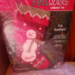 May include: Felt applique kit for a snowman stocking. The kit includes wool yarn, cotton thread, printed felt, beads, a needle, and easy instructions with an alphabet. The stocking is red with white felt accents and a snowman applique. The snowman is white with black eyes and a black mouth. The stocking has the word "Cheer" written on it. The kit is designed by Rebecca Carter.