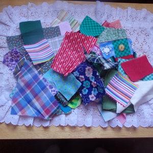 May include: A collection of colorful fabric scraps in various patterns and textures, including plaid, polka dots, stripes, and floral prints. The scraps are arranged on a white lace doily.