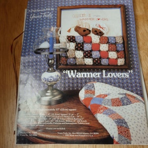 May include: A patchwork quilt kit featuring two bears in a bed with the text "Quilters make warmer lovers" and "Warmer Lovers". The kit includes fabric, batting, lace, ribbon, instructions, and a needle. The quilt is approximately 13 inches square.