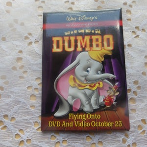 May include: A red, black, and gold pin featuring a cartoon image of Dumbo the elephant from the Disney movie Dumbo. The pin has the text "Walt Disney's 60th Anniversary Edition Dumbo Flying Onto DVD and Video October 23".