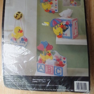 May include: A plastic canvas needlepoint kit featuring a yellow duck with a blue umbrella, a tissue box cover with a yellow duck and a pink and blue 'A' block, and a wall hanging with a yellow duck and balloons. The kit includes instructions for creating three different projects. The kit is from Bucilla and is labeled '6155'.