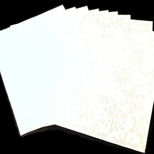 May include: A stack of off-white paper sheets, some with a snowflake pattern embossed on the surface. The paper has a slightly textured feel and is arranged against a black background. The snowflake design adds a festive touch.