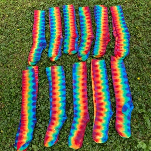 Op de afbeelding: Een verzameling van 11 kleurrijke tie-dye kniekousen. De sokken zijn in twee rijen op een bed van groen gras gerangschikt. De sokken zijn een mix van rood, oranje, geel, groen, blauw en paars.