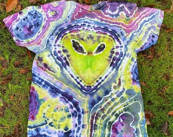 Puede incluir: Una camiseta tie-dye con un diseño de cara de extraterrestre verde en el centro. La camiseta es una mezcla de colores morado, azul, verde y amarillo.