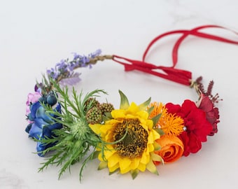 May include: A colorful flower crown with a red ribbon tie. The crown features a sunflower, orange, red, and blue flowers.