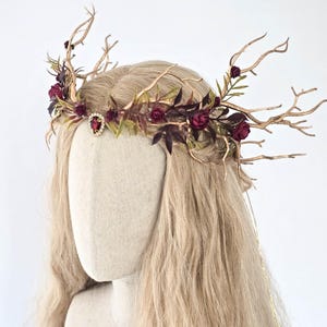 Può includere: Un copricapo artigianale con una struttura dorata a forma di ramo, ornata da rose bordeaux, foglie viola scuro e un gioiello rosso centrale. La corona è progettata per un tema fantasy o boschivo, perfetta per eventi speciali.