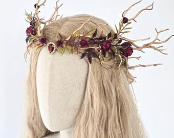 May include: A handcrafted headpiece featuring a gold-toned, twig-like frame adorned with burgundy roses, dark purple leaves, and a central red jewel. The crown is designed for a fantasy or woodland theme, perfect for special events.