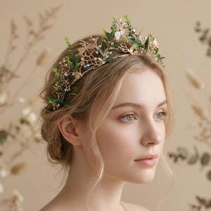 Könnte beinhalten: Ein goldfarbener Blumenkranz mit grünen Blättern und kleinen weißen Akzenten. Der Kranz wird auf dem Kopf einer Person getragen, vor einem weichen, neutralen Hintergrund. Der Kranz scheint handgefertigt zu sein und eignet sich für besondere Anlässe.