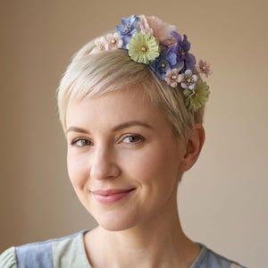 Könnte beinhalten: Ein floraler Haarreif mit pastellfarbenen Blumen, darunter rosa, blaue und grüne Blüten. Die Blumen sind auf einem dünnen, flexiblen Band angeordnet. Der Haarreif ist ein zartes Accessoire.