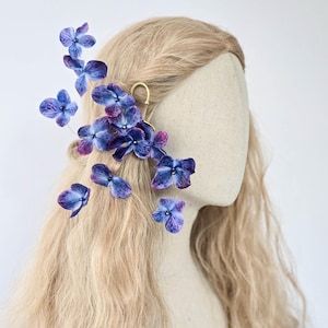 Pode incluir: Um acessório de cabelo floral com flores de hortênsia azuis e roxas. As flores são presas a um grampo de cabelo de metal dourado, projetado para ser usado no cabelo. O cabelo é longo e loiro.
