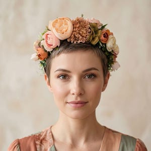 Könnte beinhalten: Eine Frau mit einem Blumenkranz aus pfirsichfarbenen, rosa- und cremefarbenen Blüten. Der Kranz sitzt auf kurzem, dunklem Haar. Die Frau trägt ein floral gemustertes Kleid in Pfirsich- und Grüntönen. Der Hintergrund ist ein neutrales Beige.