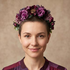 Könnte beinhalten: Ein Blumenkranz mit tiefvioletten Rosen, kleineren Blüten und Grün. Der Kranz wird auf dem Kopf einer Person getragen. Die Person trägt ein lila- und kastanienfarbenes Hemd mit Kragen. Der Hintergrund ist neutral beige.