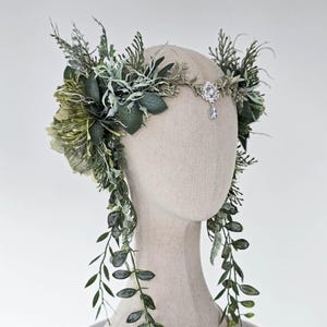 May include: A headpiece featuring an arrangement of various green leaves and foliage, including long trailing vines. The headpiece is centred with a silver-toned metal ornament containing a clear, faceted crystal drop that hangs at the forehead.