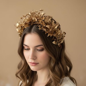 May include: A gold-colored floral crown with intricate details. The crown features various leaf and flower shapes, creating a natural, elegant design. The headpiece is worn by a person with long, wavy brown hair, against a neutral background.