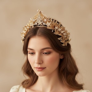 May include: A gold-colored bridal crown featuring star and floral designs, adorned with small pearls and crystals. The crown is worn by a person with long, wavy brown hair and a cream-colored dress. The background is a soft, neutral tone.