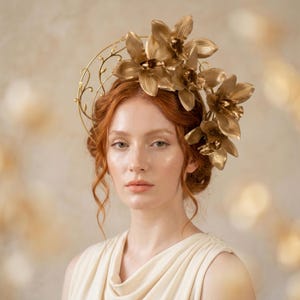 May include: A gold-toned headpiece featuring a circular wire frame adorned with delicate vine-like details and a cluster of metallic orchid flowers. The accessory is positioned atop the head, showcasing a structured botanical design in a polished finish.