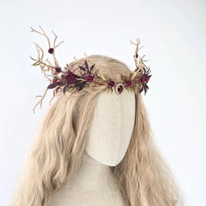 May include: A floral crown with burgundy roses, green leaves, and gold-toned branches. A small, ornate jewel is centered on the crown. The crown is displayed on a mannequin head with long, blonde hair, against a white background.
